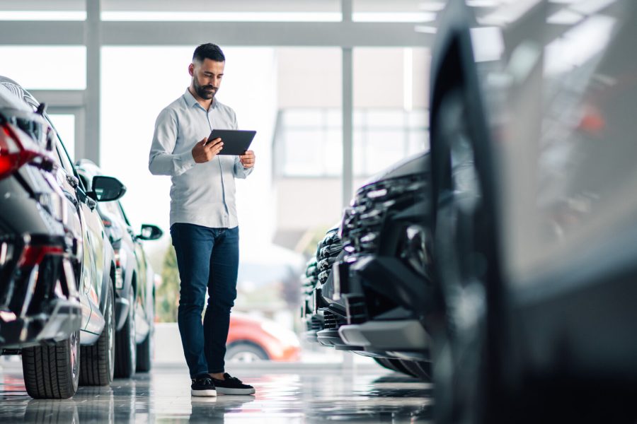 Automotive retailer using a tablet to inspect vehicle information between rows of cars in a modern showroom.